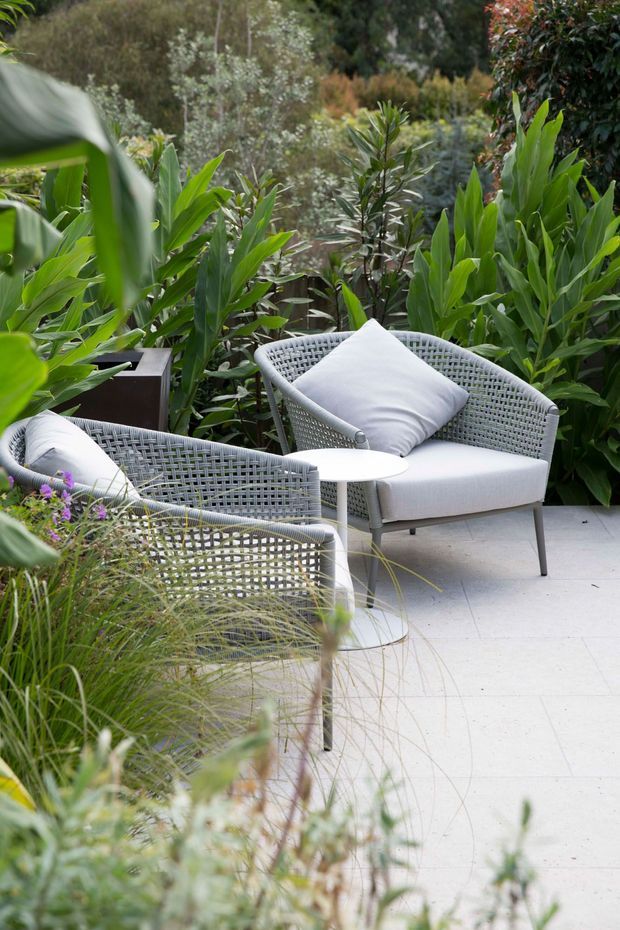 A landscaped outdoor seating area with two woven lounge chairs, pale cushions, and small round side tables arranged on a light paved terrace. Dense layered tropical and ornamental planting surrounds the furniture, creating a private garden setting.