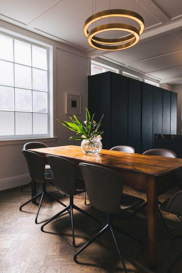 A refined dining room with a large rectangular dark timber dining table surrounded by black upholstered chairs. The room features tall frosted-paned windows, dark vertical timber cabinetry or partitioning, a warm sculptural ring pendant light, white coffered ceiling detailing, and a herringbone timber floor. A glass vase with green foliage forms the table centrepiece.