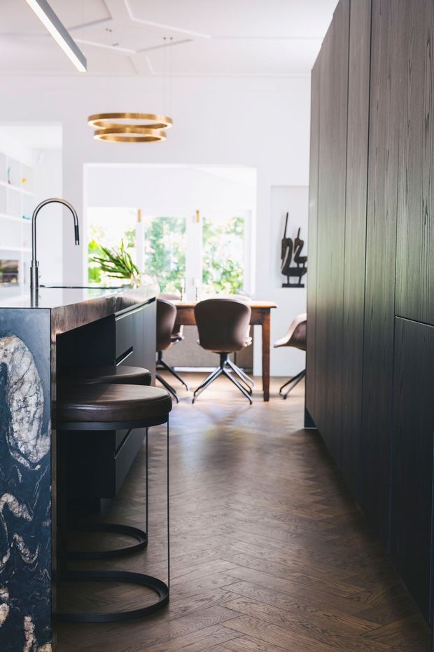 Bright, refined kitchen opening into a dining area. The kitchen features a dark stone island with curved seating, black tapware, dark vertical-panel cabinetry, and a warm herringbone timber floor. A brass ring pendant hangs above, while large windows and white walls create an airy, high-contrast interior.