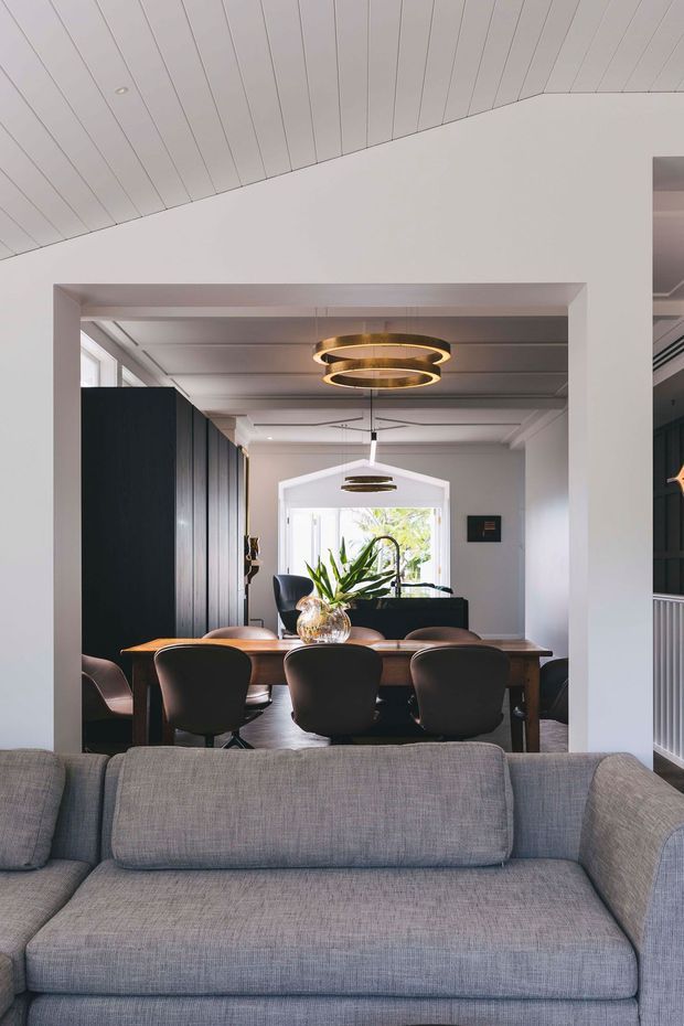 A contemporary open-plan interior viewed from behind a large grey sofa. Through a wide opening is a dining area with a dark timber dining table, several black upholstered chairs, a large indoor plant centerpiece, dark vertical wall panelling, and layered circular pendant lights. The room has white walls, a white painted timber-lined sloping ceiling, generous glazing, and a bright, refined atmosphere.