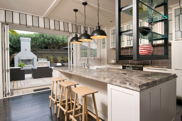 A bright, refined kitchen with a large marble-look island and waterfall-style benchtop, white cabinetry, black-framed glass display joinery, and three hanging pendant lights. Timber stools line the island, while folding or glazed openings connect the kitchen to an outdoor entertaining area with a built-in fireplace. Decorative leadlight-style glazing adds character.