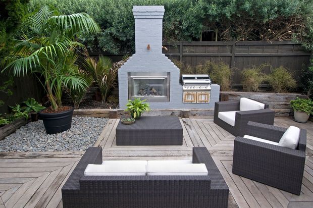 Landscaped outdoor entertaining area with a paved and timber-decked patio, dark woven outdoor lounge furniture with white cushions, a built-in white masonry fireplace, and a stainless-steel outdoor barbecue set against dense planting and a timber privacy fence.
