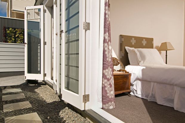 A bright bedroom opening directly onto a small gravel courtyard through fully glazed white bi-fold doors. The room contains a bed with white bedding, a patterned upholstered headboard, wooden bedside furniture, and a warm table lamp. Dark stepping stones lead across the gravel outside, with screening and neighbouring residential elements visible beyond.