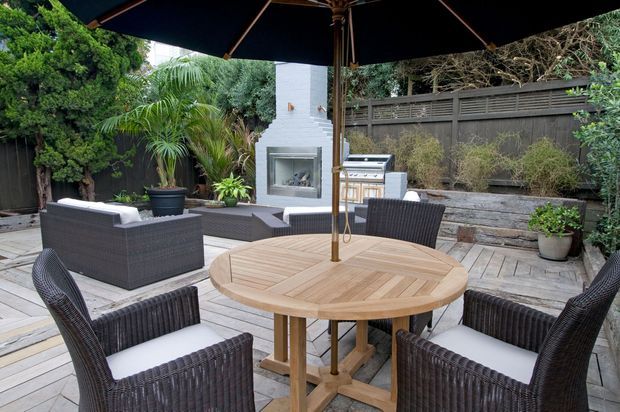 A landscaped outdoor entertaining area with a timber deck, round timber dining table, woven outdoor chairs, large umbrella, built-in barbecue, and a white rendered outdoor fireplace surrounded by planting and timber screening.