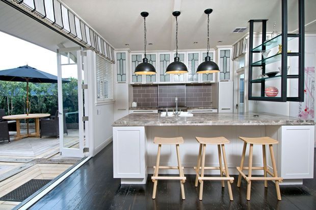 Bright, spacious kitchen with a large central island, marble-look benchtop, white cabinetry, patterned glass-front overhead cabinets, tiled splashback, and three black pendant lights. Timber stools sit at the island, while full-height glazed doors open to an outdoor deck and dining area. A built-in open shelving unit with decorative objects is visible on the right.