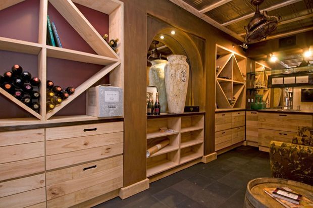 A richly detailed wine cellar and tasting room with custom timber cabinetry, geometric open wine racks, drawers, shelving, and display niches. Dark slate flooring, warm ambient lighting, exposed timber ceiling elements, and a central timber-clad island or bar create a cozy, rustic-luxe atmosphere. Wine bottles and cellar storage dominate the space.