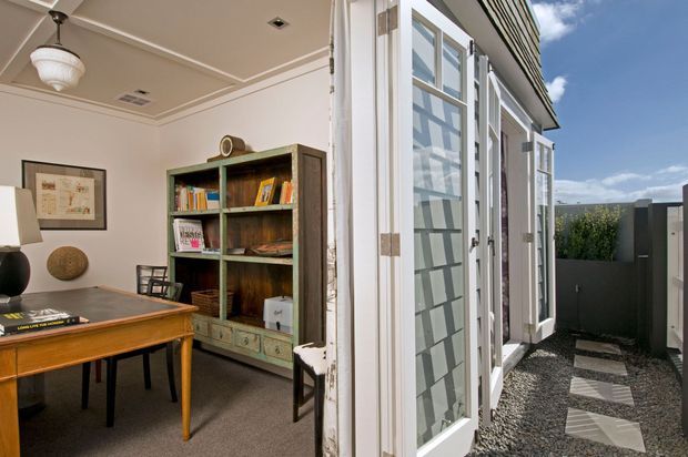 A split interior-exterior view showing a compact home office opening onto a narrow outdoor walkway. Inside are a vintage wooden desk, chairs, framed artwork, a tall distressed built-in-style bookcase, carpeted flooring, and a decorative pendant light. Outside, white-framed glazed doors and windows line the facade beside a gravel path with rectangular stepping stones and a dark privacy fence. The building has a low-pitched roof with horizontal exterior cladding.