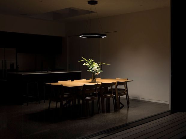 A dimly lit contemporary dining area viewed from a dark adjoining space or exterior threshold. A long light-wood dining table is surrounded by dark timber chairs and illuminated by a suspended geometric pendant light. A kitchen island and dark cabinetry are visible to the left, while a large plain wall and dark polished floor create a restrained, intimate atmosphere. A leafy floral arrangement forms the centrepiece.