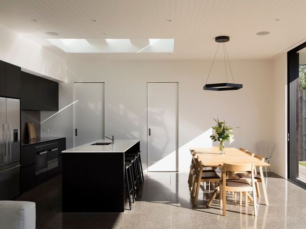 Bright contemporary open-plan kitchen and dining area with matte black cabinetry, a central island with integrated sink and seating, a timber dining table, and large glazed doors opening to the exterior. The space features a white linear ceiling, skylight, polished speckled flooring, minimalist pendant lighting, and strong natural sunlight.