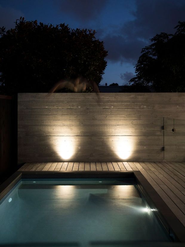 A moody nighttime view of a compact private swimming pool surrounded by timber decking. A tall horizontal timber-clad privacy wall is illuminated by two ground-mounted lights, with dense planting silhouetted against a dark blue evening sky. The pool water and integrated underwater lighting create a calm, atmospheric outdoor setting.