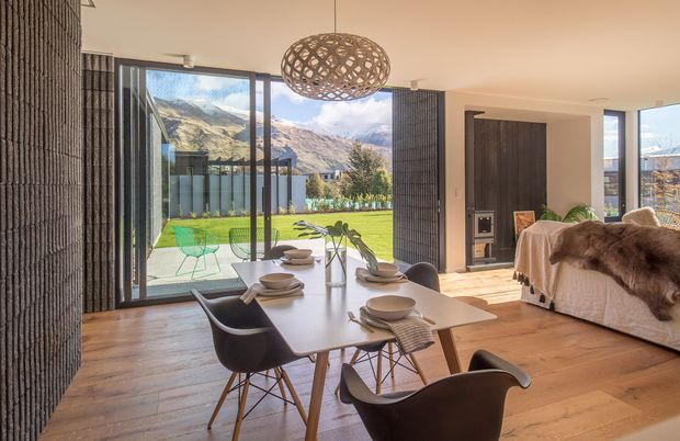 Open-plan dining and living area with a contemporary timber interior, dark textured feature walls, floor-to-ceiling glazing, and a sculptural woven pendant light. A dining table with modern chairs occupies the foreground, while a fireplace and sofa are visible beyond. The glazing opens views toward a manicured lawn and dramatic mountains.