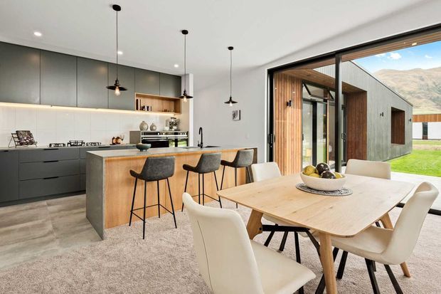 Open-plan contemporary kitchen and dining area with a large dark-grey linear kitchen, timber-clad island, integrated appliances, pendant lighting, and a light timber dining table with upholstered chairs. Full-height black-framed glazing opens toward a timber-lined outdoor courtyard and landscaped setting with mountains visible beyond. The room has a refined, warm-modern feel with strong indoor-outdoor connection.