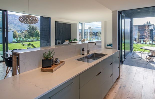 Bright, open-plan contemporary kitchen with a large pale stone island, integrated sink and dark tap, extensive minimalist cabinetry, timber flooring, and floor-to-ceiling glazing opening to a lawn and patio. A dining area and living space are visible beyond the island, with mountain scenery seen through the windows.