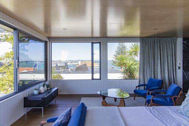 Bright coastal-style bedroom or sitting room with a bed in the foreground, blue upholstered lounge chairs, a round coffee table, dark sideboard, full-height corner glazing, and a broad harbour or coastal view. The room features vertical wall panelling, a warm timber ceiling, dark timber flooring, and layered curtains.