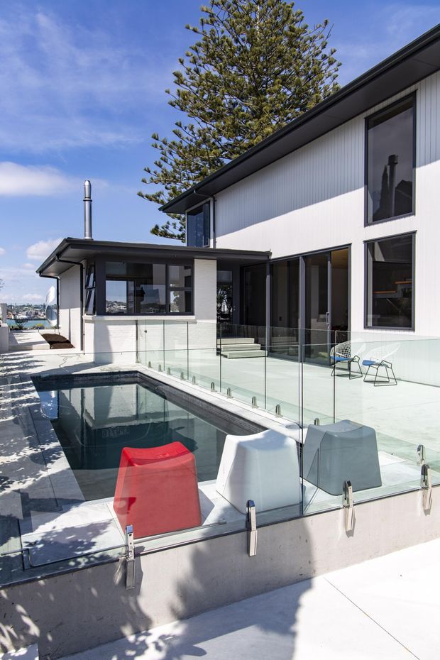 A bright contemporary residential exterior centered on a narrow swimming pool and surrounding terrace. The two-storey home has white vertical cladding, dark metal roof edges and window frames, extensive glazing, and a glass pool safety barrier. Outdoor lounge chairs sit beside the pool, with a mature conifer and a glimpse of the coastal urban setting beyond.