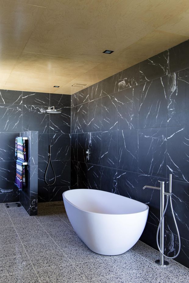 Contemporary bathroom with a freestanding white oval bathtub, wall-mounted bath filler, open shower area, dark marble-look wall tiles with prominent white veining, and grey terrazzo-style floor tiles. The ceiling is clad in warm-toned timber veneer with recessed downlights.