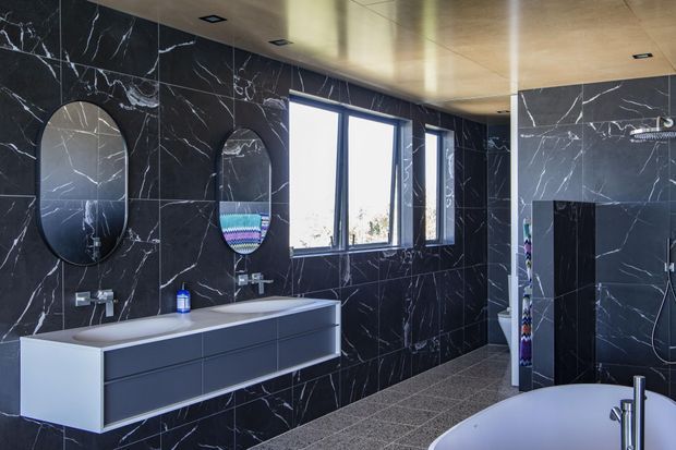 Contemporary bathroom with dramatic black marble-look wall tiles, a floating double vanity with integrated basins, oval mirrors, large windows, a walk-in shower enclosure, and a partially visible freestanding bathtub. The space has a warm timber-toned ceiling, dark fixtures and cabinetry, and a refined monochromatic palette.