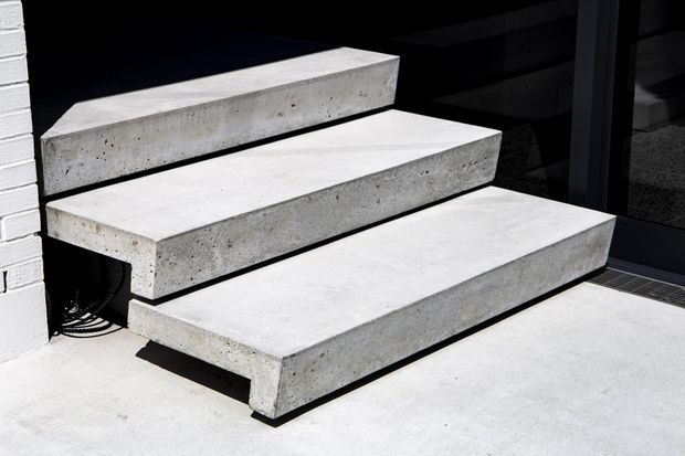 Close-up architectural photograph of three broad floating concrete steps leading to a dark-framed glazed entrance. The steps have smooth pale grey concrete surfaces with exposed aggregate and visible black shadow gaps beneath, beside a white-painted brick wall.