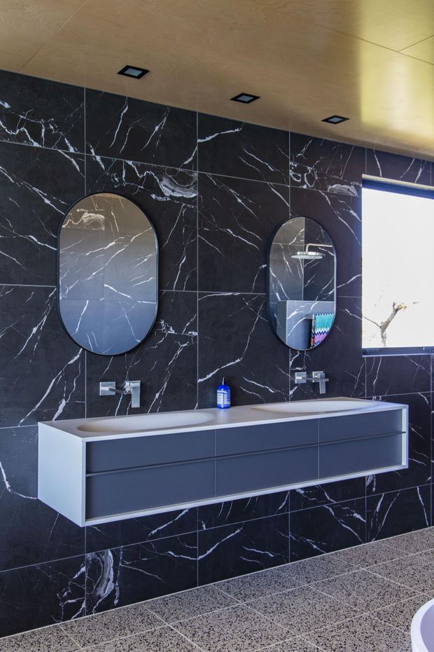 A contemporary bathroom featuring a long floating double vanity with integrated basins, dark grey cabinetry, chrome wall-mounted fixtures, and two rounded-rectangle mirrors. The walls are clad in dramatic black marble-look tiles with white veining, complemented by a warm timber-look ceiling, recessed downlights, and a speckled grey floor. A freestanding bath is partially visible at the lower right.