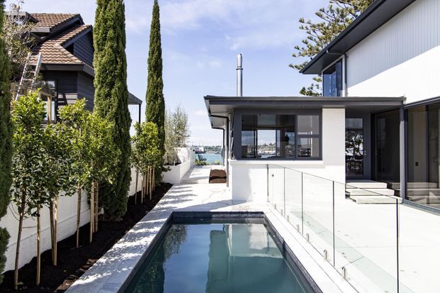 Bright exterior view of a contemporary multi-level residence with a narrow swimming pool, glass balustrades, white rendered walls, dark cladding, and a flat-roofed pavilion. Tall cypress trees and landscaped planting line the side path, while water is visible in the distance.