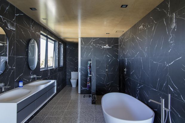 A spacious contemporary bathroom featuring dramatic dark marble-look wall tiling with white veining, a matching double vanity and oval mirrors, a freestanding oval bathtub, a separate shower area, and a toilet. The room has a warm-toned ceiling, recessed lighting, large windows, and grey terrazzo-style flooring.