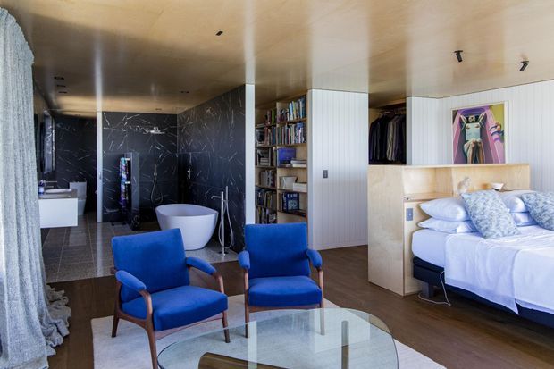 A refined open-plan bedroom suite with a freestanding bathtub and dark marble-effect bathroom wall visible behind the seating area. The bedroom features a built-in timber storage and wardrobe wall, open shelving filled with books, a large upholstered bed, blue armchairs, and a glass coffee table. The space has warm timber finishes, white bedding, blue accents, and contemporary integrated lighting.