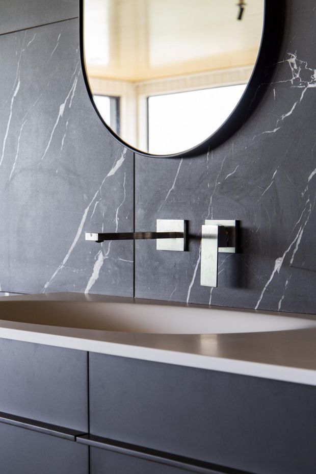 Close-up view of a contemporary bathroom vanity featuring dark charcoal marble-look wall panels with prominent white veining, a round black-framed mirror, wall-mounted brushed metal tapware, and dark built-in vanity cabinetry with a pale benchtop.