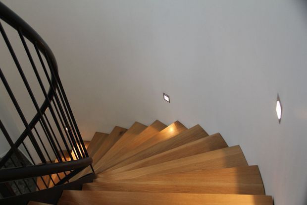 Close-up view looking down a curved staircase with warm timber wedge-shaped treads, a dark metal balustrade, and smooth light-coloured walls. Small recessed wall lights illuminate the stairwell, creating a contemporary, sculptural interior.