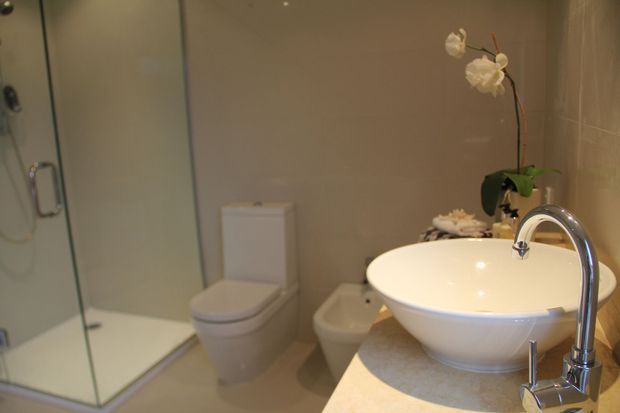 A warm, modern bathroom featuring a frameless glass shower enclosure, wall-mounted toilet, bidet, and a white vessel basin on a light stone vanity with a polished chrome gooseneck tap. Beige wall tiles and orchid accents create a calm, refined atmosphere.