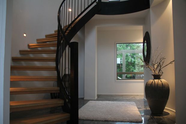 A refined interior stairway with floating timber treads, a dark sculptural curved metal balustrade, and a landing framed by white walls. A large decorative vase and branches sit beside a window, while a pale rug rests on a dark stone or tile floor.