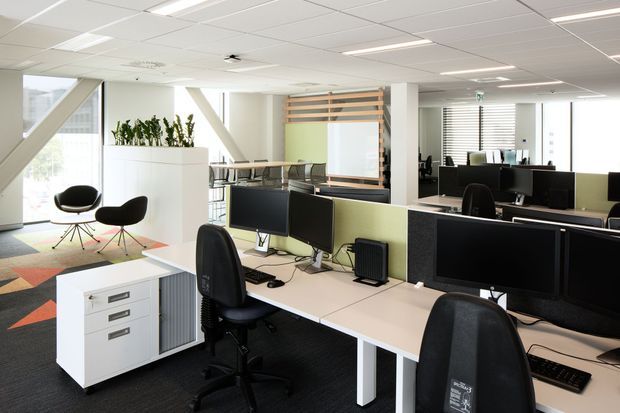 Bright open-plan commercial office with rows of workstations, large computer monitors, white desks, black office chairs, low green and black desk partitions, and a white storage pedestal. The space has a suspended acoustic ceiling with recessed fluorescent lighting, extensive glazing with horizontal blinds, white structural columns, and a colourful geometric carpet. A small informal seating area and planted divider are visible near the windows.