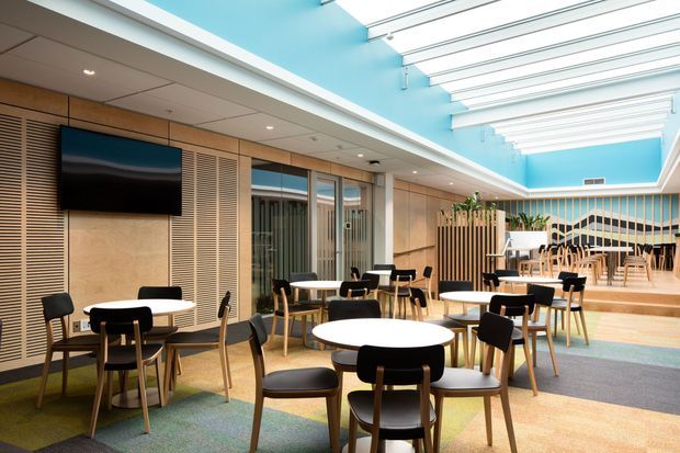 Bright contemporary commercial café or hospitality interior with a spacious dining area, round white tables, black and timber chairs, wood-toned slatted wall panelling, integrated bench seating, and a raised dining zone at the rear. A vivid turquoise ceiling and extensive linear skylight structure create an airy, daylight-filled atmosphere. A wall-mounted television and glass-partitioned areas are also visible.