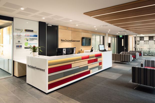 A contemporary government office reception area with a large white reception desk featuring a geometric inset front in red, grey, beige, and wood-toned panels. The space includes a timber-look wall behind the desk with New Zealand Government signage, black doors, glazed office partitions, patterned carpet tiles, tiled flooring, striped lounge seating, indoor plants, recessed lighting, and a linear timber slat ceiling feature.