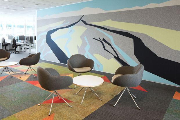 Bright contemporary office breakout lounge with sculptural upholstered lounge chairs arranged around small round white tables. A large abstract geometric mural spans the feature wall, while open-plan workstations are visible behind a glazed partition. Multicoloured geometric carpet tiles and a white acoustic ceiling complete the vibrant collaborative space.
