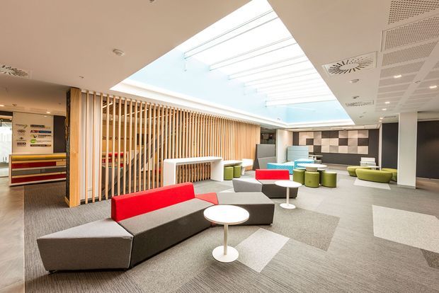 A bright contemporary commercial lounge or reception area with modular grey seating, vivid red and lime-green upholstered accents, small round tables, and a large central skylight. A timber vertical-slat screen separates the seating zone from the circulation area, while patterned acoustic ceiling panels and a geometric wall feature add visual interest. Carpet tiles cover the floor throughout.