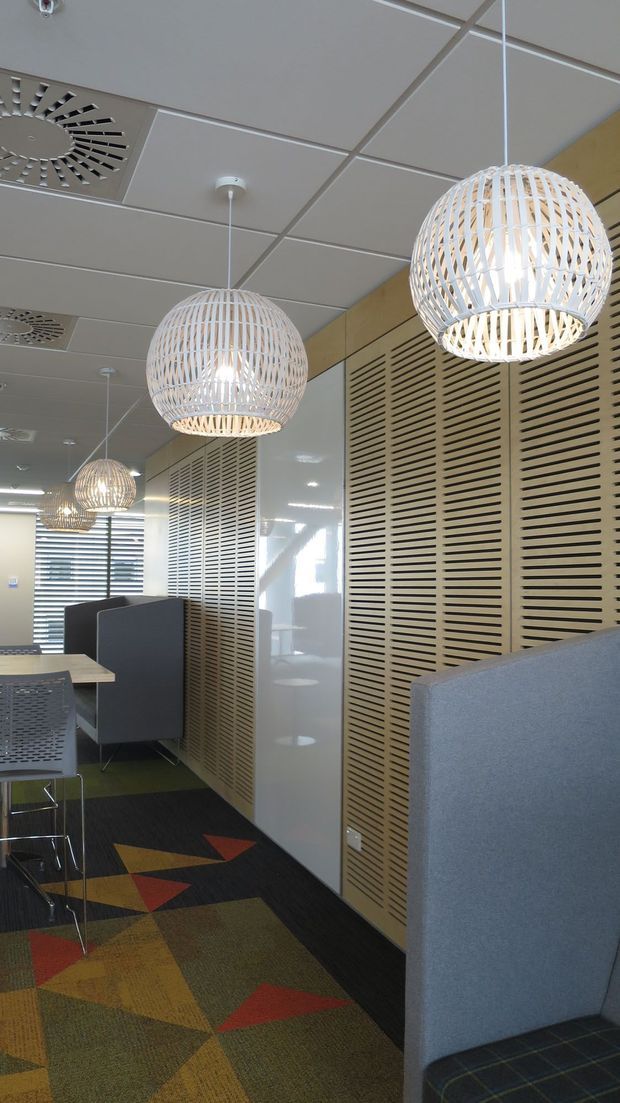 A contemporary commercial office interior with acoustic privacy screens, booth-style seating, patterned carpet tiles, a suspended grid ceiling, and multiple woven spherical pendant lights. The space uses warm timber-toned slatted partitions alongside grey upholstered furniture and colourful geometric flooring.
