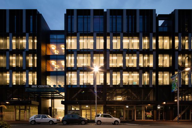 Night-time exterior photograph of a contemporary multi-storey commercial building with a dark structured facade, extensive illuminated curtain-wall glazing, vertical fins, recessed balconies or terraces, and active ground-floor retail or office frontage. Streetlights, signage, and parked cars are visible along the foreground.