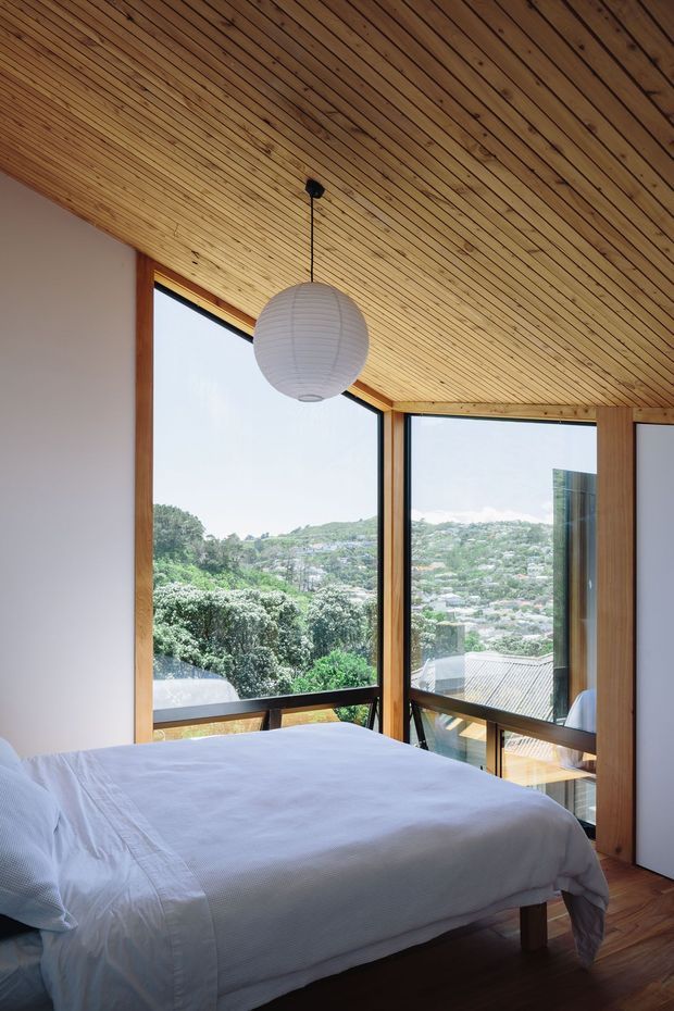 A bright bedroom with a low-sloped exposed timber-lined ceiling, white bedding, and large floor-to-ceiling corner glazing framed in warm timber. The windows overlook a leafy hilly suburban landscape, while a simple white spherical pendant provides a minimal focal point.