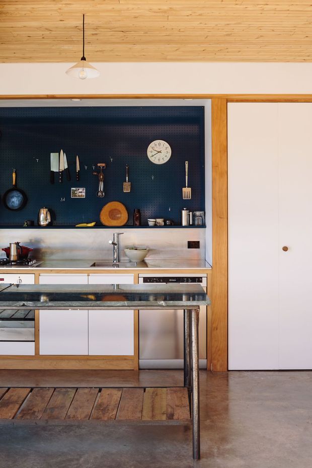 A compact, carefully detailed kitchen with warm timber ceiling and framing, white flat-panel cabinetry, a dark stone island benchtop, stainless-steel appliances and tapware, and a prominent deep blue pegboard splashback displaying cookware and utensils. A white pendant light hangs above, while a large white door is integrated into the timber wall framing. The room combines practical open storage with a warm, workshop-like character.