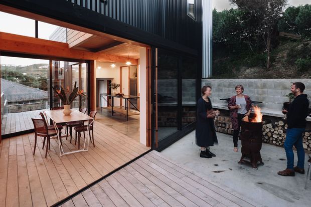 A contemporary multi-level home with a large open timber deck and glazed indoor-outdoor living area. The dining space has a timber table and chairs, exposed timber ceiling and beams, and extensive floor-to-ceiling glazing. Adjacent to the dark corrugated-clad exterior wall is a concrete-paved courtyard with a freestanding outdoor fire pit, stacked firewood, and several people gathered around it. The setting appears elevated and wooded, with a view across a hilly landscape.