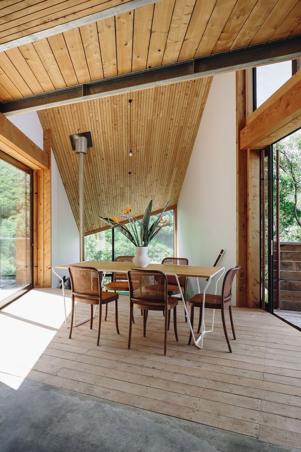 Sunlit dining area in a timber-framed pavilion or cabin, featuring a long rectangular table with woven cane dining chairs, extensive floor-to-ceiling glazing, exposed timber post-and-beam construction, and a dramatic sloping timber-lined ceiling. Natural vegetation is visible outside.