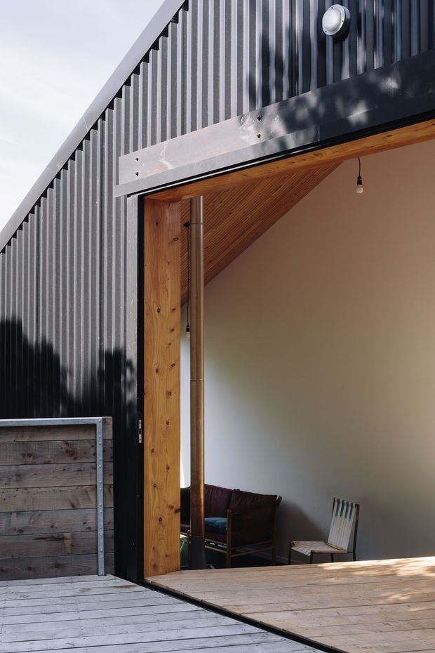 A close architectural view of a modern dark corrugated-metal-clad building opening onto a timber deck. The large open glazed or garage-style opening reveals a warm timber-lined sloping ceiling, exposed timber framing, a compact seating area, and a simple pendant light. The scene combines black ribbed metal, natural timber, pale interior walls, and strong sunlight and shadows.