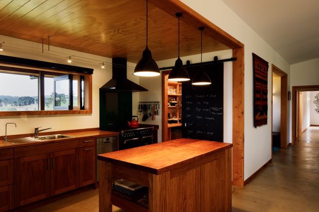 Warm, rustic-modern kitchen with extensive timber cabinetry, a large timber island, stainless-steel appliances, a farmhouse-style sink, and multiple black pendant lights. The room opens to a hallway and includes a timber-lined ceiling, exposed timber-framed opening, a dark rangehood, and a chalkboard pantry or storage area.