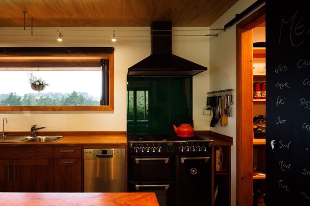 Warm, compact kitchen with dark timber cabinetry, a timber-lined ceiling, a stainless-steel dishwasher, wood-framed window, and a large black range cooker with hood. A pantry or storage space with open shelving is visible through the doorway on the right, alongside a black chalkboard wall.