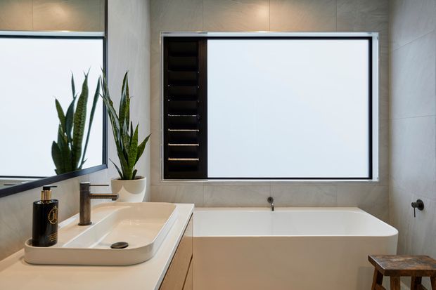 Contemporary bathroom featuring a freestanding rectangular bathtub beneath a large frosted or opaque window with a dark louvred section, a wall-mounted basin with brushed metal tapware, light stone-look wall tiles, a large corner mirror, and potted snake plants. A small wooden stool sits beside the bath.