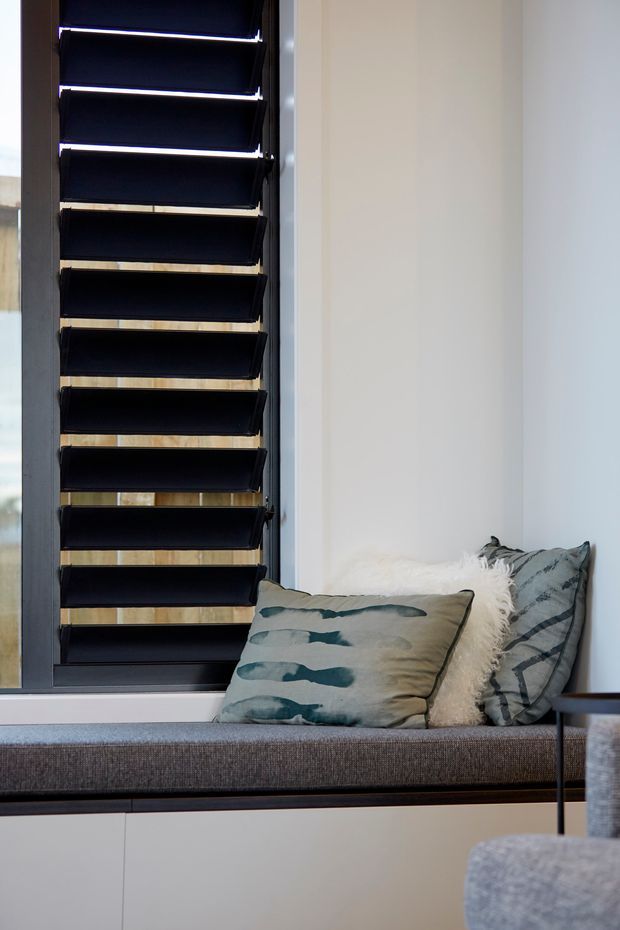 A compact built-in window seat beneath a tall louvred window, with dark horizontal blinds, pale walls, grey upholstered bench seating, and muted blue-grey patterned cushions. The space has a clean, contemporary feel with integrated joinery and natural light.
