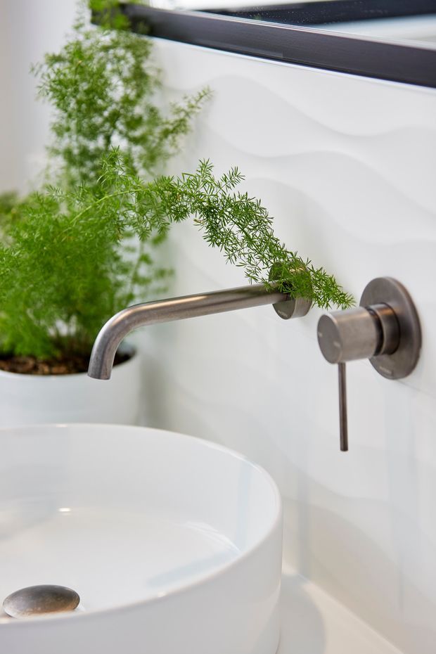 Close-up of a contemporary bathroom vanity featuring a white round vessel basin, wall-mounted brushed metal tapware, white sculptural wall tiles, and a leafy potted plant. A dark-framed mirror is partially visible above.