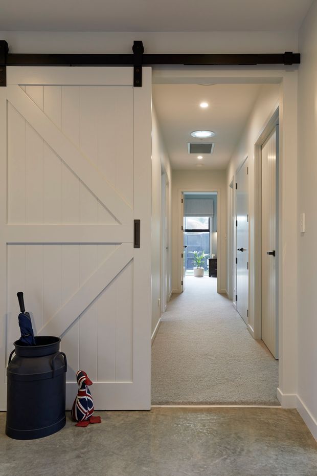 Interior view of a bright residential hallway opening from a concrete-floored entry area. A white barn-style sliding door with dark metal hardware dominates the left side, while the carpeted corridor leads to a glazed exterior door and several white interior doors. Recessed ceiling lights and a skylight-like ceiling feature illuminate the space; a small umbrella stand and decorative item sit near the entry.