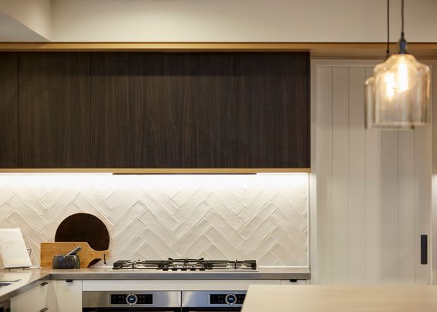Close-up of a refined contemporary kitchen, featuring dark timber overhead cabinetry, a white herringbone tile splashback, integrated stainless-steel cooktop and appliances, pale shaker-style cabinetry, and a warm glass pendant light.