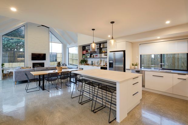 Bright open-plan kitchen and living area with a large white island, integrated seating, pendant lights, stainless-steel appliances, white cabinetry, open shelving, polished concrete flooring, and expansive glazed openings. A dining table and lounge area sit beside the kitchen, with a contemporary linear fireplace and television wall visible in the background.